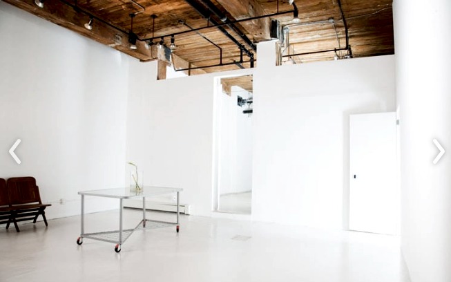 photo of the gallery space: White-walled gallery room with wooden-beam ceiling, rolling table and two wooden chairs (screenshot of a website slideshow; arrow buttons are part of the screenshot).